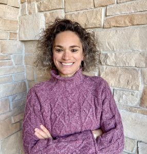headshot of Rhonda, a light-skinned woman with dark ombre curly hair, standing in front of a brick wall. She is smiling, arms crossed, and wearing a purple turtleneck sweater.