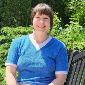 A headshot of Brandice, a white woman with straight black-brown hair tied back with bangs loose, wearing a bright blue sweater with white accents, smiling and sitting on a bench outside in front of lush green foliage.