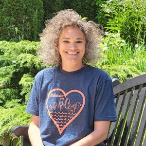 A headshot of Rhonda, a white woman with a halo of curly hair, wearing a heather blue-gray T-shirt with a coral Milwaukee decal on it, smiling and sitting on a bench outside in front of lush green foliage.