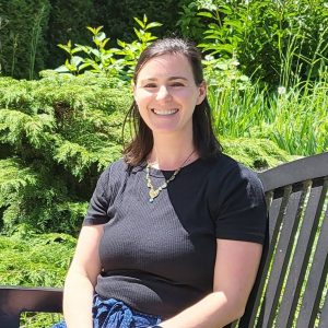 A headshot of Sami, a white woman with straight black-brown hair half-up and half-down, wearing a colorful necklace and a black T-shirt, smiling and sitting on a bench outside in front of lush green foliage.