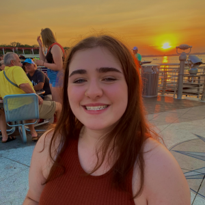 A photograph of a smiling young woman with long, straight brown hair. She sits at a restaurant table outdoors and smiles at the camera. The sunset is behind her.