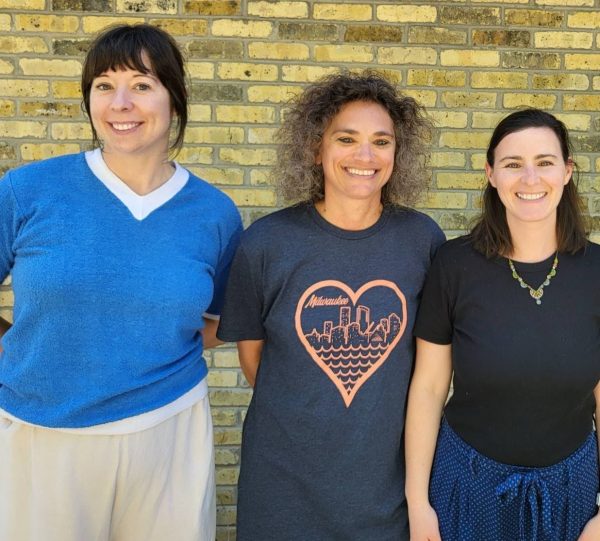 Three women standing together in a line, smiling in front of a brown brick wall. Brandice is on the far left, her dark hair tied back and wearing a blue sweater. Rhonda is in the middle left, wearing a heather blue-gray and coral Milwaukee shirt. In the middle right is Sami with a gold-tone necklace, black T-shirt, and her hair half-up, half-down.